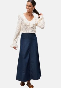 Kantblouse in crèmekleur met gerimpelde halslijn en klokmouwtjes, gecombineerd met een lange donkere spijkerrok. Bruine lederen schoenen.
