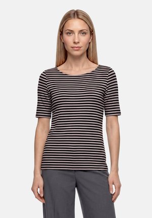 Femme aux cheveux blonds raides portant un t-shirt rayé noir et gris à manches courtes et un pantalon gris, debout devant un fond clair uni.
