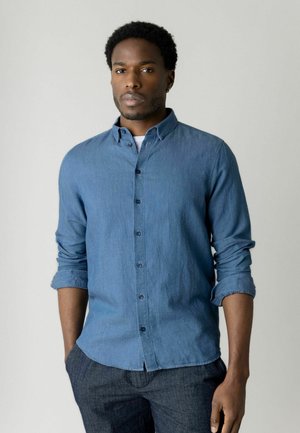 Muž s krátkymi kučeravými vlasmi, oblečený v modrej košeli s dlhým rukávom vyhrnutým hore a tmavých nohaviciach, stojaci pred jednoduchým svetlým pozadím.