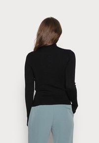 Femme aux longs cheveux bruns portant un haut noir à manches longues et un pantalon bleu clair, debout de dos devant un fond uni.