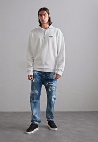 Witte zip-up sweatshirt met een hoge kraag, gecombineerd met versleten blauwe jeans met scheuren. Zwarte sneakers maken de outfit af.