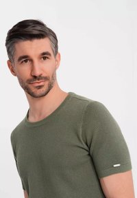 Zelený svetřík s krátkým rukávem vyrobený z texturované tkaniny, s kulatým výstřihem a malým kovovým detailem na rukávu.