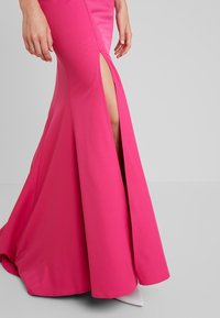 Femme portant une jupe longue rose vif avec une fente haute sur la cuisse, laissant apparaître la peau et une chaussure blanche pointue.