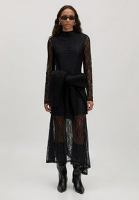 Zwarte kanten jurk met lange mouwen en een hoge hals, voorzien van een geknoopte gebreide riem in de taille. Gekleed met kniehoog zwarte laarzen.