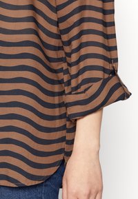 Blusa a righe marroni e blu con maniche arrotolate, realizzata in un tessuto morbido, con orlo curvo e dettagli di ventilazione laterale.