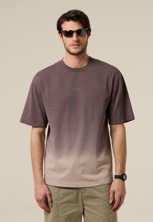 Man met zonnebril, een T-shirt met een verloop van bruin naar beige, beige broek en een zwarte polshorloge tegen een effen beige achtergrond.