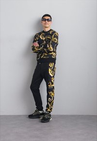 Melnā treniņtērps ar zelta spirālveida rakstiem, kas ietver kreklu ar apaļu kakla izgriezumu un sašaurinātas bikses, apvienots ar melniem sporta apaviem un saulesbrillēm.