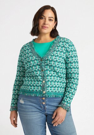 Cardigan turchese con motivo e frange, chiusura a bottoni e maniche lunghe. Indossato sopra un top verde teal, abbinato a jeans con bottoni davanti.