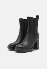 Botas de tobillo de cuero negro con tacón grueso, paneles laterales elásticos, textura suave y detalles de costura mínimos. Suela de plataforma negra.