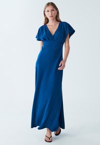 Dlhé modré šaty s výstrihom do V, krátkymi roztrasenými rukávmi, priliehavým živôtikom, splývavou sukňou a bočným rozparkom. Hladká tkanina s matným povrchom.