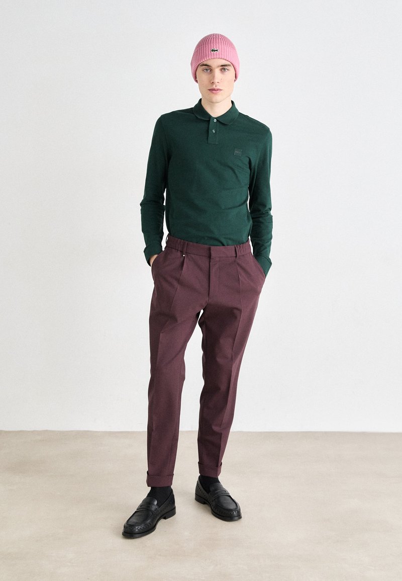 Zielona koszulka polo z długim rękawem i kieszonką na piersi, zestawiona z bordowymi, zwężającymi się spodniami i czarnymi loafersami. Różowa wełniana czapka dopełnia całość.