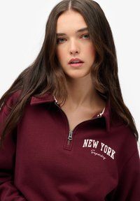 Burgundowa bluza z zamkiem błyskawicznym, z białym napisem "NEW YORK" oraz logo, z wysokim kołnierzem i miękkim, teksturowanym materiałem.