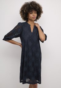 Sötétkék csipke ruha virágmintával, V-kivágással és rövid puffos ujjakkal. Bonyolult részletek díszítik a szegélyt.