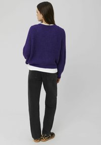 Pull violet oversize en maille avec une finition texturée, superposé à une chemise blanche, associé à un jean noir droit et des chaussures à motifs.