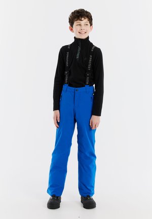 Blauwe sneeuwbroek met verstelbare zwarte bretels, hoge taille en zijzakken. Draagt een zwart langemouwenshirt en zwarte schoenen.