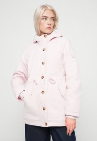 Jovem com cabelo loiro curto veste uma jaqueta com capuz rosa claro, com grandes botões e as mãos nos bolsos, olhando para a direita.