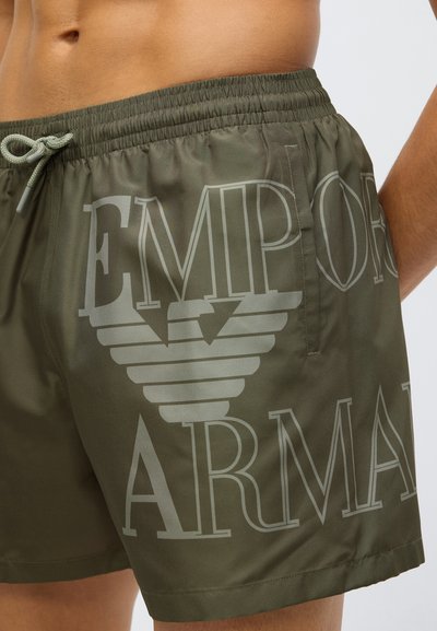 Olíva zöld fürdőnadrág rugalmas derékpánttal, húzózsinórral és nagy, világosszürke "EMPORIO ARMANI" logóval. Sima, könnyű anyagból.