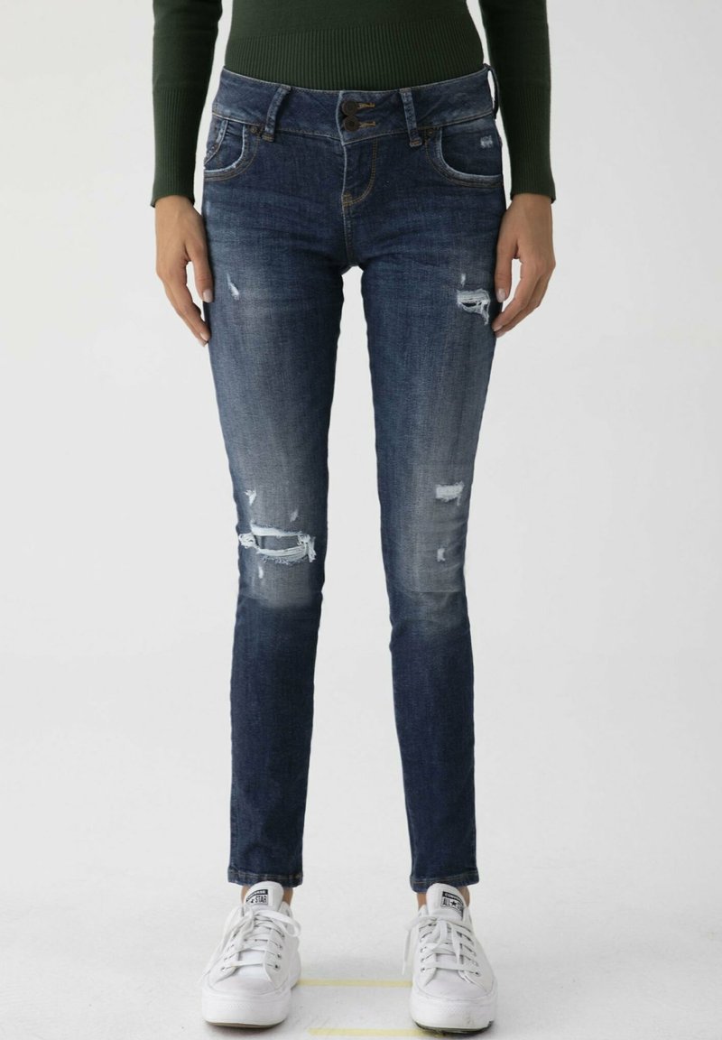 Jean skinny en denim bleu foncé, présentant des patchs usés, de petites abrasions et une coupe ajustée. Associé à des baskets blanches.