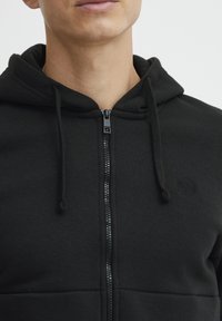 Sudadera negra con cremallera hecha de tela suave. Cuenta con una capucha ajustable, un frente con cremallera y un logo texturizado en el pecho.