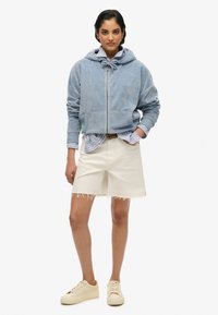 Sudadera de forro polar azul claro con cremallera, camiseta blanca de rayas debajo y pantalones cortos blancos con dobladillo deshilachado. Completado con zapatillas crema.