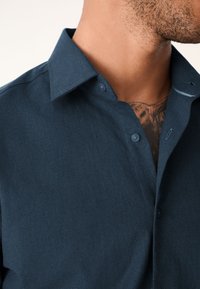 Marineblauw overhemd met knopen en een effen textuur, voorzien van een puntige kraag en een enkele knoop op de kraag, close-up van het bovenste kledingstuk.