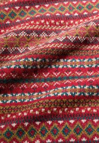 Adītā tekstila audumam ir atkārtota ģeometriska raksta kompozīcija sarkanā, zaļā, zilā un baltā krāsā, ar mīkstu tekstūru un nelielām biezuma variācijām.