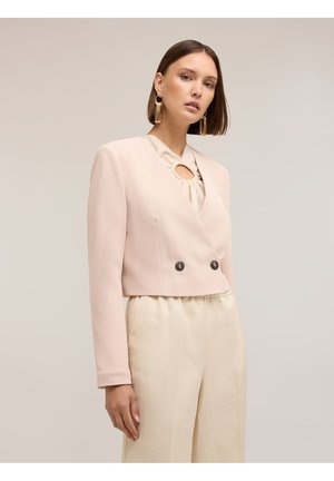 Kvinde med kort brunt hår iført beige cropped blazer med to knapper over cremefarvede satinbukser og store gyldne hængende øreringe.