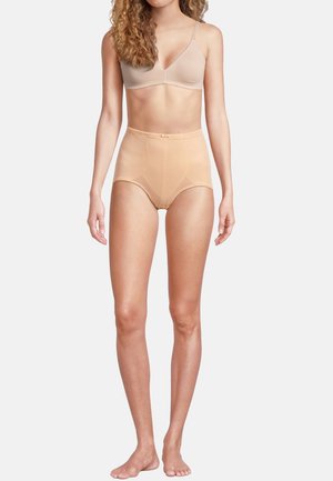 Kvinde iført beige højtaljede formende trusser og en nude bh, stående barfodet mod en ens hvid baggrund.