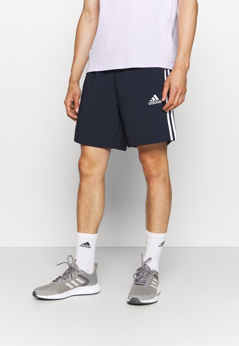 Hombre con pantalones cortos deportivos Adidas color azul marino con rayas blancas, calcetines blancos de Adidas y zapatillas de correr Adidas grises, de pie contra un fondo blanco.