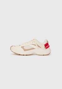 Sneaker crema e beige con tomaia in rete texturizzata, suola in gomma e branding rosso sul tallone. Presenta lacci e un design a pannelli a contrasto.