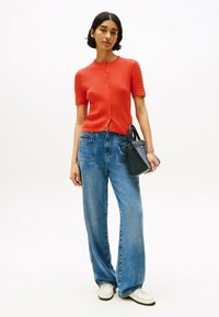 Rote strukturierte Strickjacke mit kurzen Ärmeln, knöpfbar, kombiniert mit hoch taillierten blauen weit geschnittenen Jeans. Kleine schwarze Handtasche und weiße Schuhe.