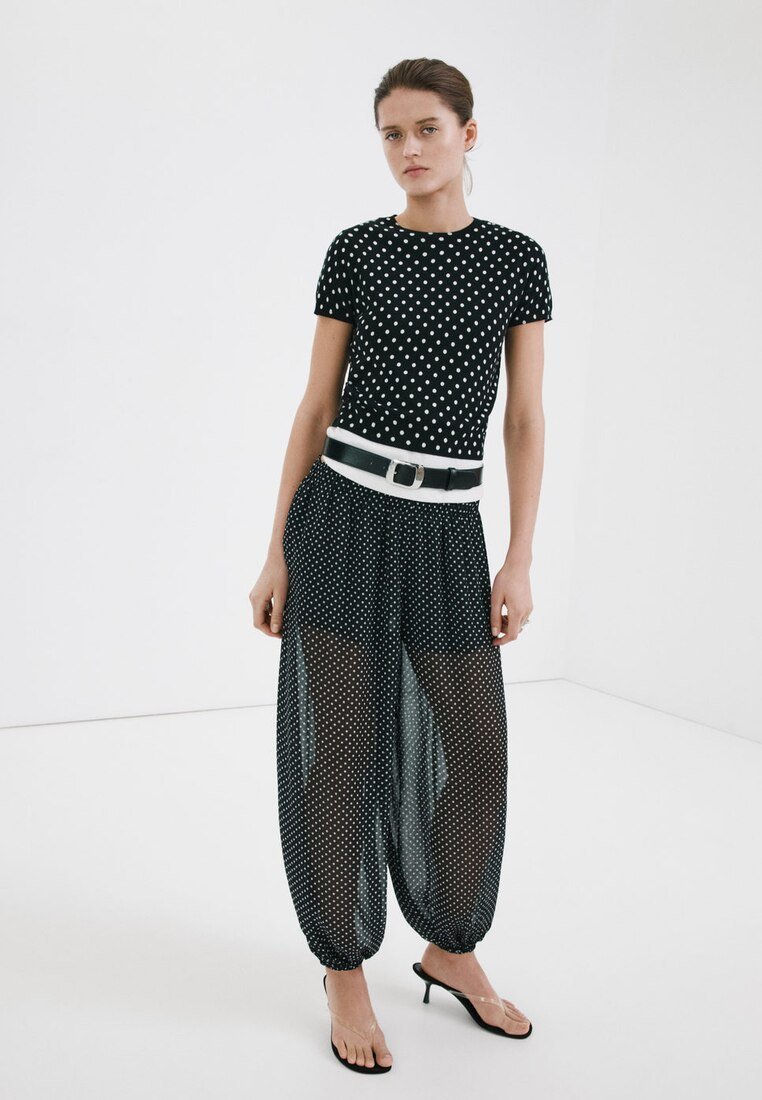 Femme debout portant un haut noir à pois manches courtes et un pantalon transparent à pois assorti avec une ceinture blanche et noire et des talons noirs.