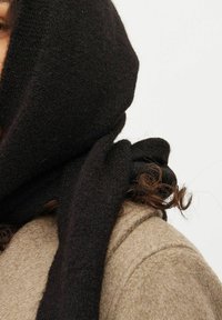 En sort uldhalstørklæde lægger sig omkring en beige uldfrakke, som fremhæver en blød tekstur og en knude detalje på siden, der delvist dækker halsen.