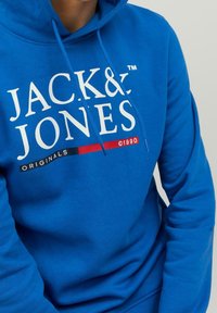 Osoba v jasně modré mikině s kapucí, na které je bílý nápis "JACK & JONES ORIGINALS ©1990" a červený a černý design na hrudi.