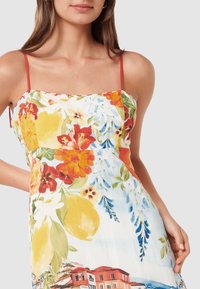 Γυναίκα που φοράει φόρεμα χωρίς μανίκια με λεπτούς κόκκινους ιμάντες και πολύχρωμο σχέδιο με λουλούδια και λεμόνια, κρατώντας το ένα χέρι πίσω από την πλάτη της.