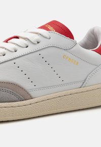Hvite lær sneakers med røde detaljer, grå semsket skinn, perforeringer på siden, gullmerke og en strukturert beige yttersåle.