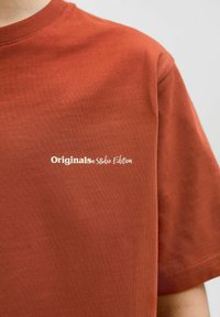Rūsas krāsas īsām piedurknēm T-krekls, izgatavots no gluda kokvilnas auduma, ar baltu logo tekstu "Originals Studio Edition" uz priekšpuses.