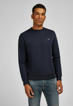 Navy Crewneck-Pullover aus weichem Material, mit gerippten Bündchen und Saum, sowie einem kleinen Logo auf der Brust. Getragen mit hellblauen Jeans.
