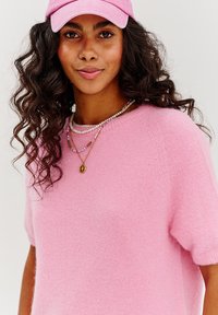 Svetloružový sveter s krátkymi rukávmi, z jemnej látky. Viacvrstvové náhrdelníky s korálkami a zlatým príveskom. Ružová čiapka. Kučeravé vlasy.