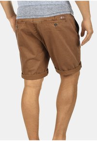 Pantalones cortos de algodón marrón con dobladillos enrollados, que presentan bolsillos traseros con cierres de botón, textura suave y un ajuste entallado.