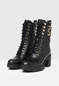 Schwarze Leder-Ankle-Boots mit hohem Blockabsatz, Schnürung vorne, goldenen Ösen und einem seitlichen Reißverschluss; mit einem metallenen Schnallenakzent.