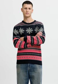 Maglione a maglia nera con motivi di fiocchi di neve e geometrici in rosso e bianco. Maniche e orlo a coste. Modello con indosso jeans, braccia incrociate.