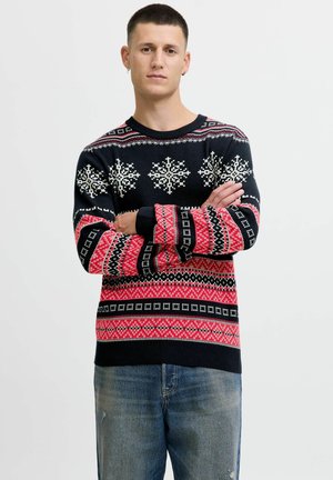 Suéter negro de punto con patrones de copos de nieve y geométricos en rojo y blanco. Mangas y dobladillo acanalados. Modelo con jeans, con los brazos cruzados.