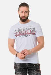 Weißes T-Shirt mit grafischem Druck in Rot und Schwarz, runder Ausschnitt, kurze Ärmel und taillierter Schnitt. Jeans mit Reißverschlusstaschen und Akzenten.