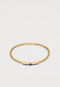 Guldtonet kædearmbånd med en rektangulær sort og guld spænde indgraveret med "GUESS" på en hvid baggrund.