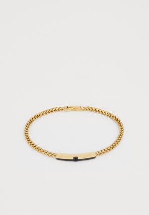 Bracelet en chaîne à maillons curb doré, avec un fermoir rectangulaire noir et doré gravé "GUESS" sur fond blanc.