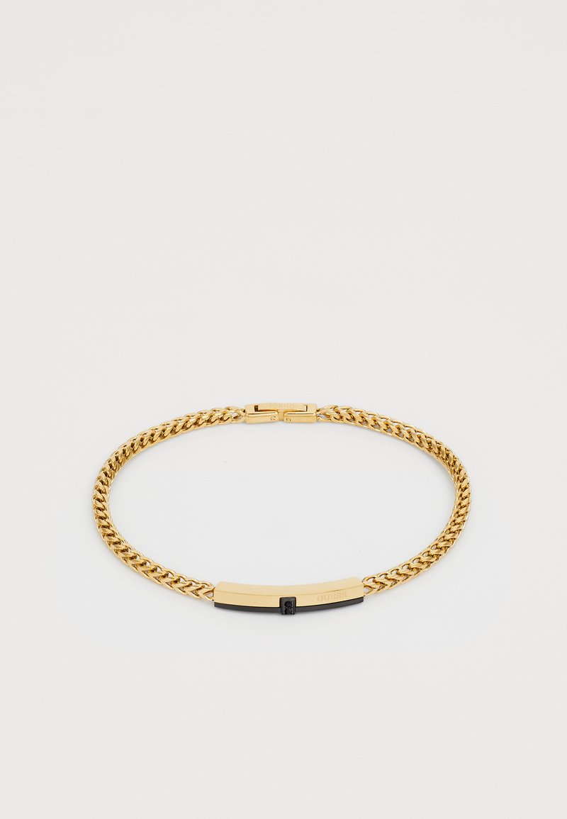 Guldtonet kædearmbånd med en rektangulær sort og guld spænde indgraveret med "GUESS" på en hvid baggrund.
