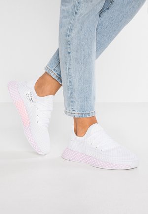 Osoba w jasnoniebieskich dżinsach i białych trampkach z różowymi, wzorzystymi podeszwami, stojąca na białej podłodze na tle białej ściany.