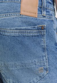 Lichtblauwe denim jeans met een achterzak voorzien van een metalen knoop en een reliëf leren label in bruin met de tekst "NO EXCESS." Subtiele textuur en stiksels zichtbaar.