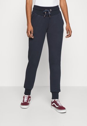Persoon draagt donkerblauwe joggerbroek met een trekkoord in de taille en bordeauxrode en witte sneakers, staand op een lichte vloer.